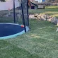 A round trampoline with a basketball hoop sits in a backyard with green grass and rock landscaping.